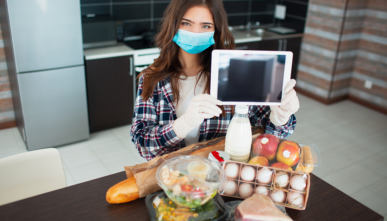 Food delivery concept. A young woman in a medical mask and a rubber disposable gloves orders food using a tablet at home. On the table are milk, salads in boxes, meat, food, fruits, eggs, bread,