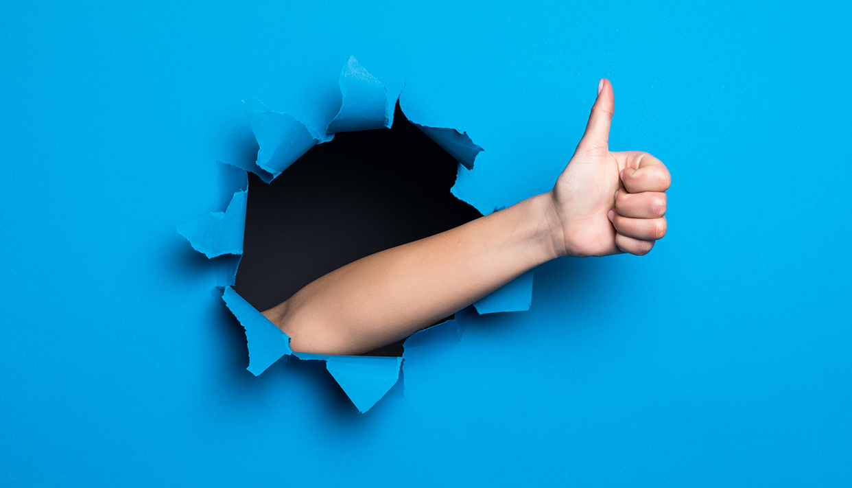 Close up of woman hand with thumbs up gesture through blue hole in paper wall.