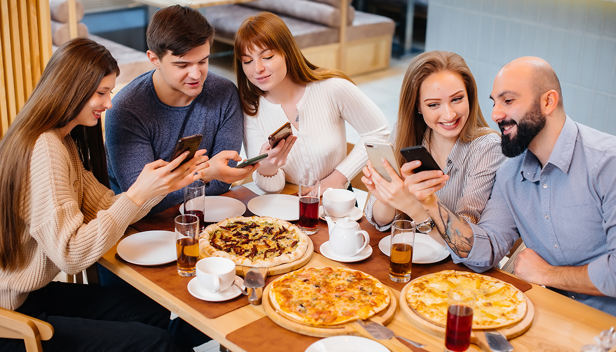 A group of young cheerful friends is sitting in a cafe talking and taking selfies on the phone. Lunch at the pizzeria